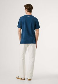 Camiseta azul marino de manga corta con un detalle de logo en la parte trasera, combinada con pantalones blancos y zapatos de color claro. Diseño simple y casual.