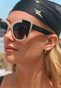 Mustad ja nude värvi ülepaisutatud kassipositiivsed prillid tumedate läätsedega. Raamil on käe peal brändi logo ja seda täiendab must peapael.