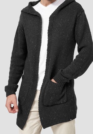 Homme portant un cardigan à capuche tricoté gris foncé sur un t-shirt blanc et un pantalon beige, une main dans la poche.