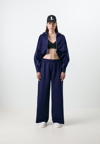 Casaco cropped azul-marinho com fecho, sutiã desportivo preto e calças largas azul-marinho. Modelo usa um gorro preto com um emblema branco de 'L'. Ténis brancos.