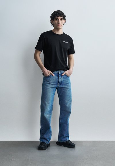 Νεαρός άνδρας στέκεται μπροστά σε λευκό τοίχο, φορώντας μαύρη μπλούζα, μπλε τζιν τζιν και μαύρα παπούτσια, με τα χέρια στις τσέπες.