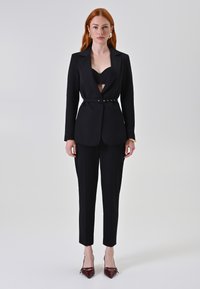 Completo sartoriale nero con giacca aderente dotata di cintura in vita, dettaglio a taglio e pantaloni a sigaretta coordinati; indossato con décolleté appuntiti color bordeaux.