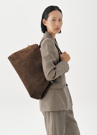 Mujer joven con traje color topo que lleva un gran bolso tote de ante marrón sobre el hombro, vista de perfil sobre un fondo blanco liso.