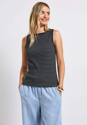 Blandet kvinde med blond hår, der smiler, iført en sort og hvid stribet ærmeløs top, lyseblå denimbukser og et stort guldbelagt armbånd.