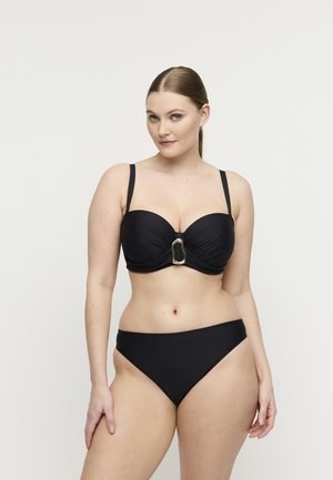 Vrouw met lichte huid en bruin haar in een zwart bikini met gouden details op de top, staand tegen een effen witte achtergrond.