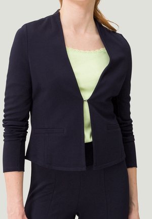 Femme portant un blazer bleu marine ajusté sur un haut vert clair orné de dentelle, avec un pantalon bleu marine assorti.