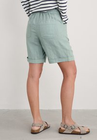 Svetle zelené lněné šortky s volným střihem, s ohrnutými manžetami a dvěma zadními kapsami. Kombinované se šedými sandály s křížovým popruhem.