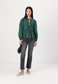 Blusa peplum verde com um detalhe em forma de chave na frente e laço, combinada com jeans retos cinza e sapatilhas bege, em uma modelo.
