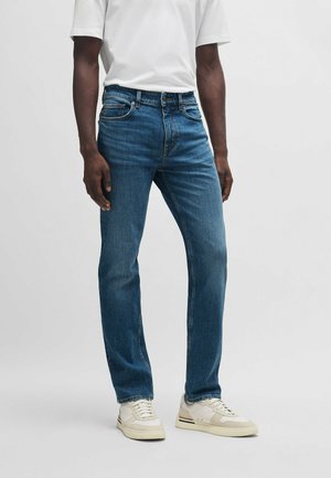 Jeans de mezclilla azul oscuro con un corte recto, diseño de cinco bolsillos, trabillas para cinturón y una textura ligeramente desgastada. Combinados con una camiseta blanca y zapatillas.