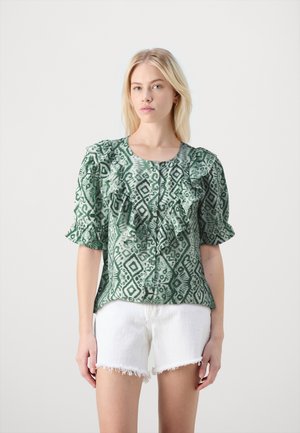 Blouse verte à motif avec des volants aux manches et sur le devant. Le tissu présente des motifs géométriques en vert plus foncé. Portée avec un short blanc effiloché.