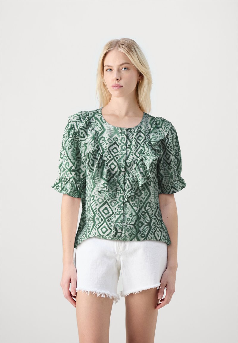 Blusa verde a fantasia con volant sulle maniche e sul davanti. Il tessuto presenta motivi geometrici in verde scuro. Indossata con pantaloncini bianchi sfilacciati.