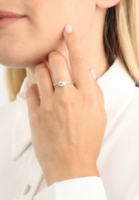 Anello d'argento con un centro in perla e accenti a forma di foglia, montato su una fascia liscia. Mano che tocca delicatamente la guancia, sfondo di colore neutro.