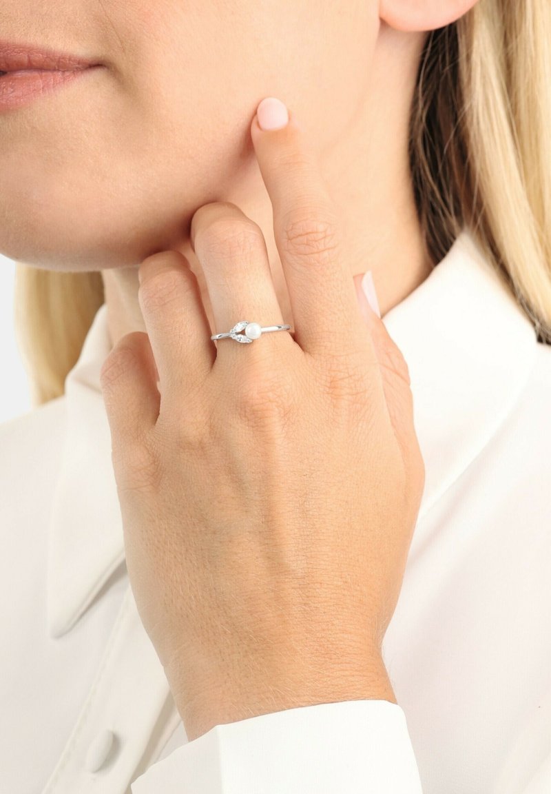 Anello d'argento con un centro in perla e accenti a forma di foglia, montato su una fascia liscia. Mano che tocca delicatamente la guancia, sfondo di colore neutro.