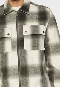 Chaqueta de cuadros hecha de una mezcla de lana en verde y crema. Cuenta con un cierre de cremallera, dos bolsillos en el pecho con botones a presión y un cuello.