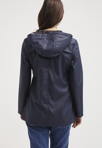 Kvinna med långt brunt hår iförd en marinblå vattentät jacka med huva och blå jeans, sedd bakifrån mot en neutral bakgrund.