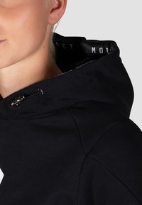Schwarze Kapuzenjacke mit strukturierter Oberfläche, ausgestattet mit silbernen Elementen und einem bedruckten Logo am Innenkragen. Nahaufnahme von Schulter und Gesicht.