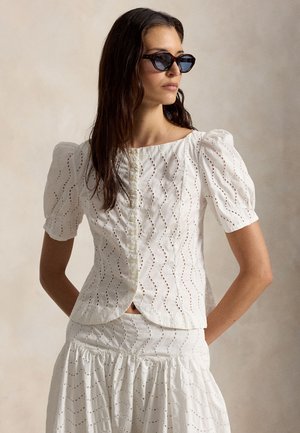 Žena v bielej blúzke s balónovými rukávmi z vyšívanej látky s gombíkmi a zodpovedajúcej sukni, doplnená čiernymi slnečnými okuliarmi v tvare mačacích očí, otočená mierne doprava.