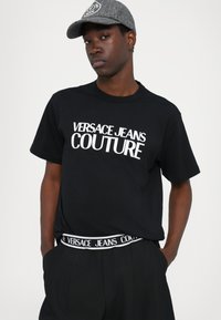 Juodas marškinėliai su balta "VERSACE JEANS COUTURE" užrašu, derinami su juodomis kelnėmis su prekiniu juosmeniu. Pilkos medžiagos kepurė.