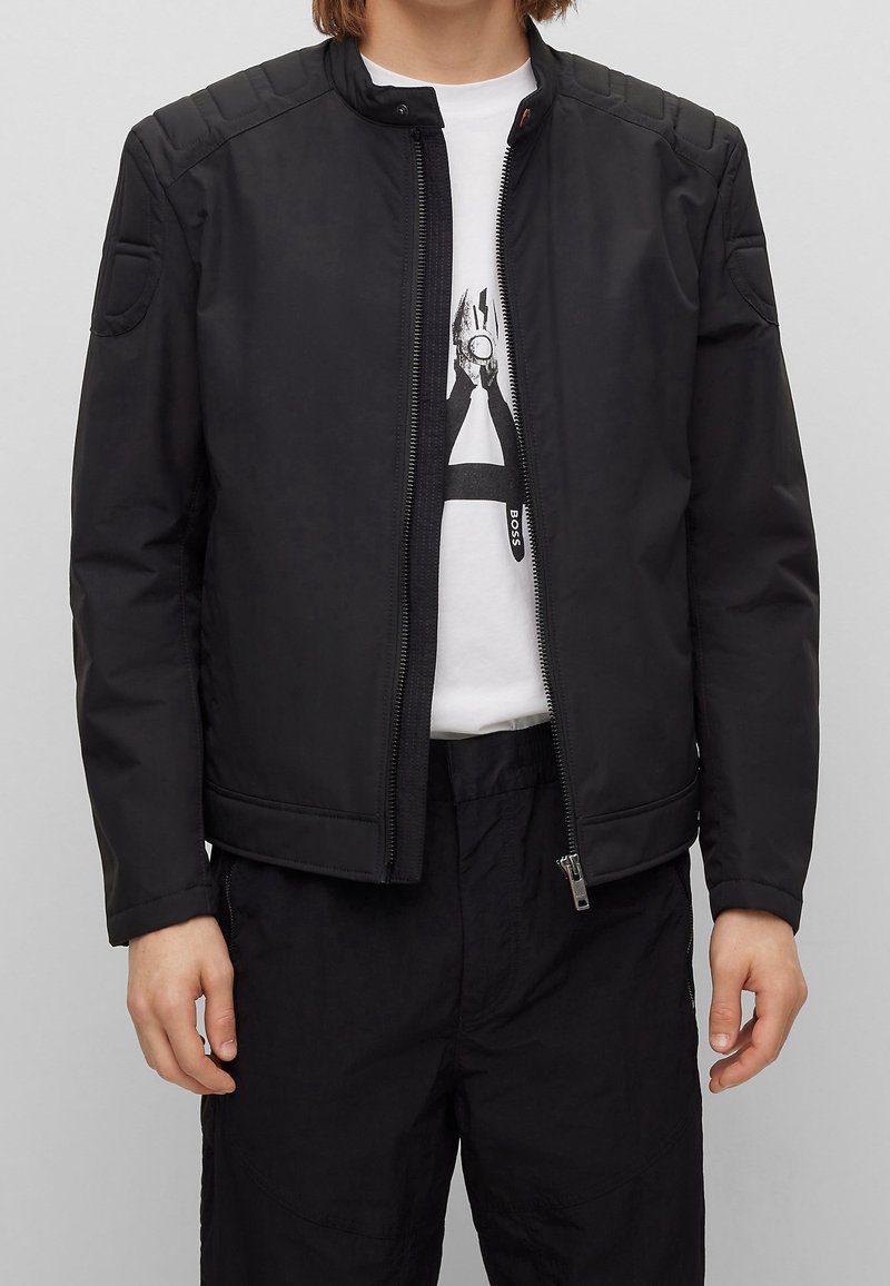 Veste bomber noire avec une fermeture éclair à l'avant, des épaules rembourrées et des détails de col côtelé. Associée à un t-shirt blanc et un pantalon noir.