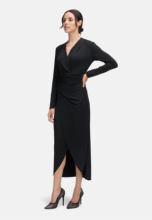 Zwarte lange mouwen jurk met een aansluitende silhouet, geplooide voorkant en een hoge split, met subtiele sparkles verspreid over de stof. Gecombineerd met zwarte hoge hakken.