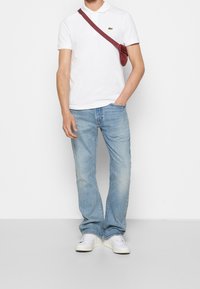 Polo blanc à manches courtes, jean droit bleu clair, et baskets blanches. Petit logo vert sur le polo. Sac bandoulière bordeaux.