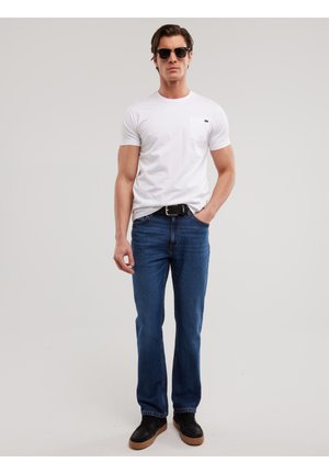 Vīrietis melnās saulesbrillēs, baltā T-kreklā, zilās džinsās, melnā jostā un melnos sporta apavos ar brūnām zolēm stāv ar vienu roku kabatā.