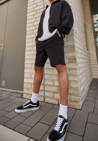 Sweat à capuche noir zippé sur un t-shirt blanc, short noir et baskets Vans noires avec des accents blancs et des chaussettes, posés devant un mur de briques.