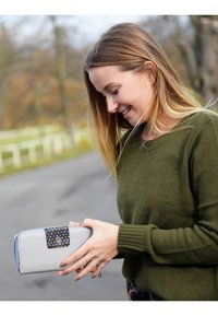 Gray wallet with a blue zipper and a black flap featuring colorful dots, held in hands. The background shows a blurred natural setting.