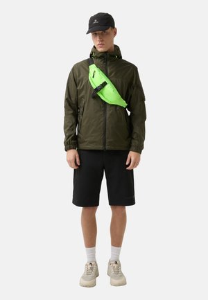 Olivenfarvet letvægtsjakke med lynlås, parret med sorte shorts. Har en lys grøn crossover-taske og sneakers i neutrale farver.