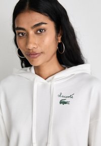 Hanorac alb cu snur și logo-ul brodat Lacoste verde. Textură netedă, cu o croială lejeră. Cercei rotunzi argintii.