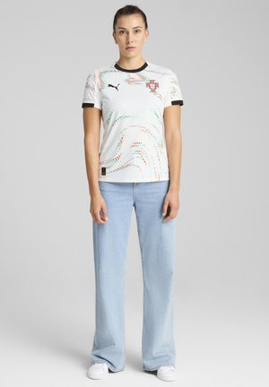 White sports jersey with red and green dot patterns, black collar and sleeves, featuring a logo on the chest. Worn with light blue jeans.