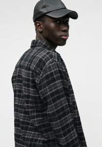 Μαύρο και γκρι καρώ φανέλα πουκάμισο με γιακά, φορεμένο κάτω από ένα μαύρο δερμάτινο καπέλο. Το πουκάμισο διαθέτει απαλό υλικό και μακριά μανίκια.