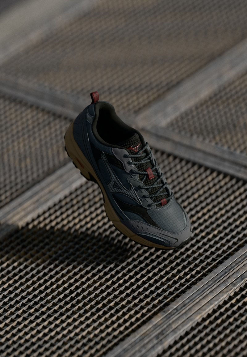 Pantofii sport de culoare gri închis și negru, cu talpă bej și accente roșii, așezați pe un podea din grilaj metalic.