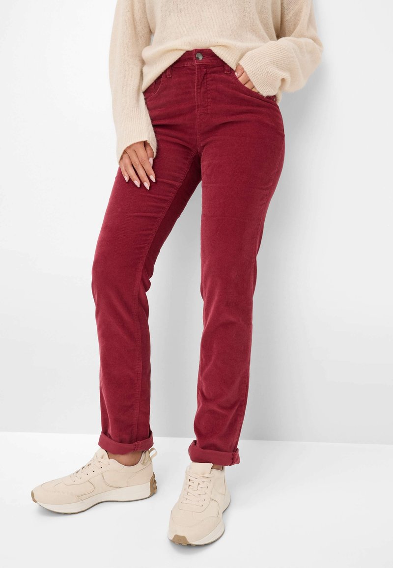 Femme portant un pantalon en velours côtelé rouge retroussé aux chevilles, un pull beige et des baskets beiges, debout devant un fond blanc.
