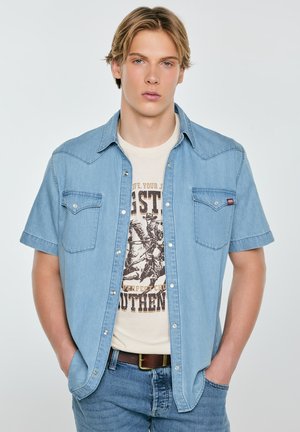 Młody mężczyzna w jasnoniebieskiej, krótko rękawowej koszuli jeansowej na beżowej koszulce z nadrukiem, z rękami w kieszeniach dżinsów na jednolitym tle.