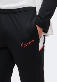Ръка в джоба, носеща черен панталон Nike с червен логотип Swoosh, комбиниран с черен и бял дълъг ръкав.