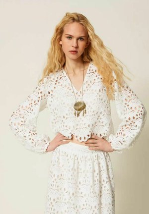 Donna bionda con capelli lunghi che indossa un top bianco in pizzo a maniche lunghe e gonna, mani sui fianchi e collana con ciondolo a forma di conchiglia.