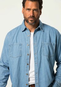 Camicia in denim azzurro chiaro con maniche lunghe, chiusura con bottoni sul davanti, due tasche sul petto e colletto classico. Abbinata a una t-shirt bianca sotto.