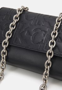 Borsa a tracolla in pelle nera con motivi geometrici in rilievo e una robusta tracolla in catena argentata su uno sfondo bianco.