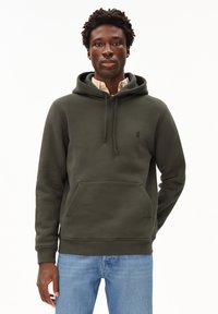 Felpa verde oliva realizzata in tessuto morbido, con tasca frontale e coulisse, con un piccolo logo sul petto. Indossata sopra una camicia a quadri.