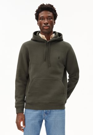 Sweat à capuche vert olive en tissu doux, avec une poche avant et un cordon de serrage, portant un petit logo sur la poitrine. Porté par-dessus une chemise à carreaux.