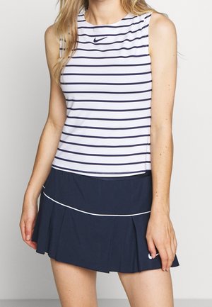 Vrouw draagt een wit mouwloos topje met zwarte horizontale strepen en een marineblauwe geplooide rok met witte biesdetails.
