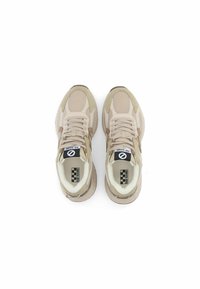 Sneaker beige con materiali in mesh e suede, forma arrotondata, suola bianca e lacci prominenti. Branding visibile sulla linguetta.