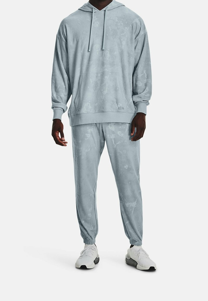 Homme portant un sweat à capuche tie-dye bleu clair et un pantalon de jogging assorti, avec des baskets blanches, debout sur fond blanc.