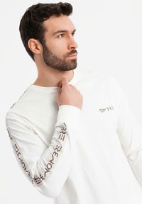 Muž s tmavými vlasy a vousy, oblečený v bílé košili s dlouhým rukávem, na které jsou hnědý text na rukávech a na hrudi, hledí do strany, ruka u krku.