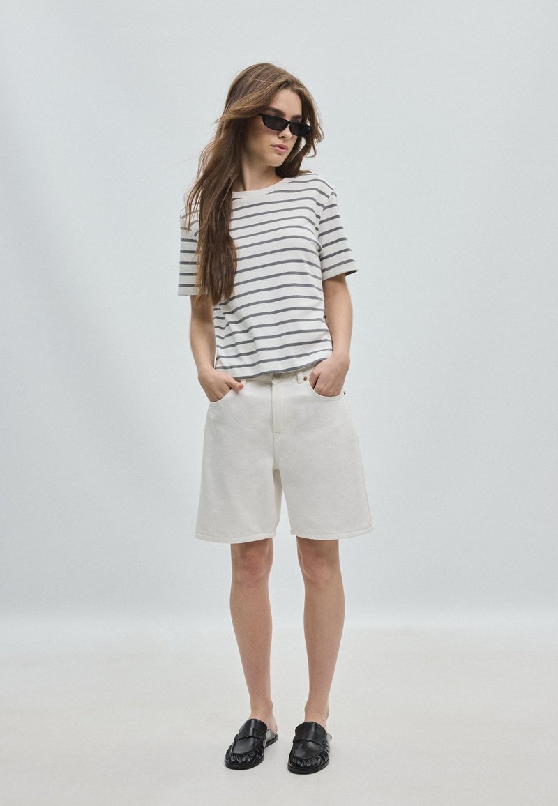 Femme portant un t-shirt rayé à manches courtes, un short blanc jusqu'aux genoux, des chaussures noires sans lacets et des lunettes de soleil noires, debout les mains dans les poches.