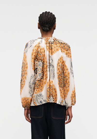 Femme aux cheveux tressés portant un chemisier à manches longues avec des motifs floraux orange, noir et blanc, vue de dos sur un fond uni.