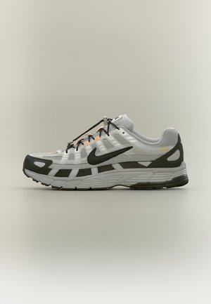 Baskets blanches et grises avec le swoosh Nike noir, lacets noirs et accents orange sur un fond neutre.