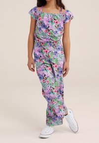 Floral jumpsuit in purple and green, featuring short puff sleeves, smocked waist, and a mix of tropical patterns. Paired with white sneakers.