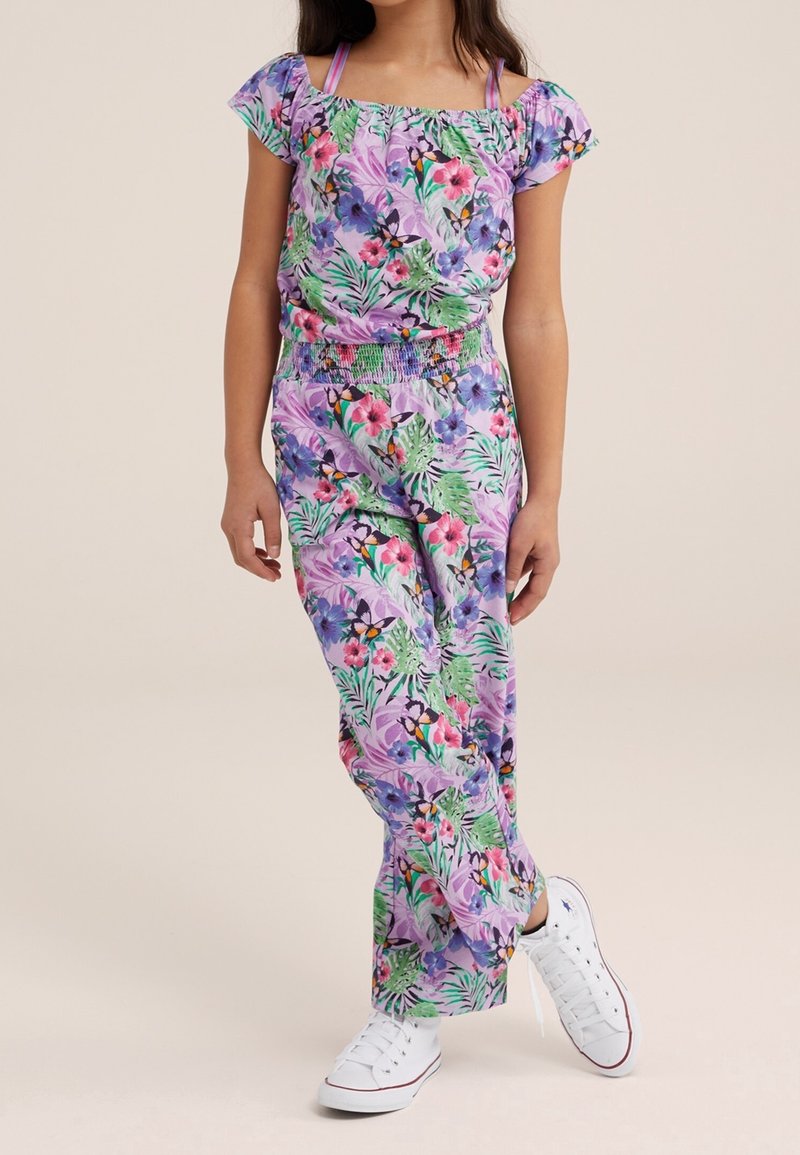 Floral jumpsuit in purple and green, featuring short puff sleeves, smocked waist, and a mix of tropical patterns. Paired with white sneakers.